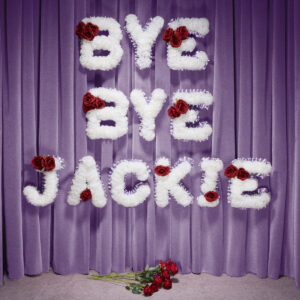 Weiße Blumenbuchstaben mit roten Rosen formen die Worte „Bye Bye Jackie“ vor einem lilafarbenen Vorhang, darunter ein Strauß roter Rosen.