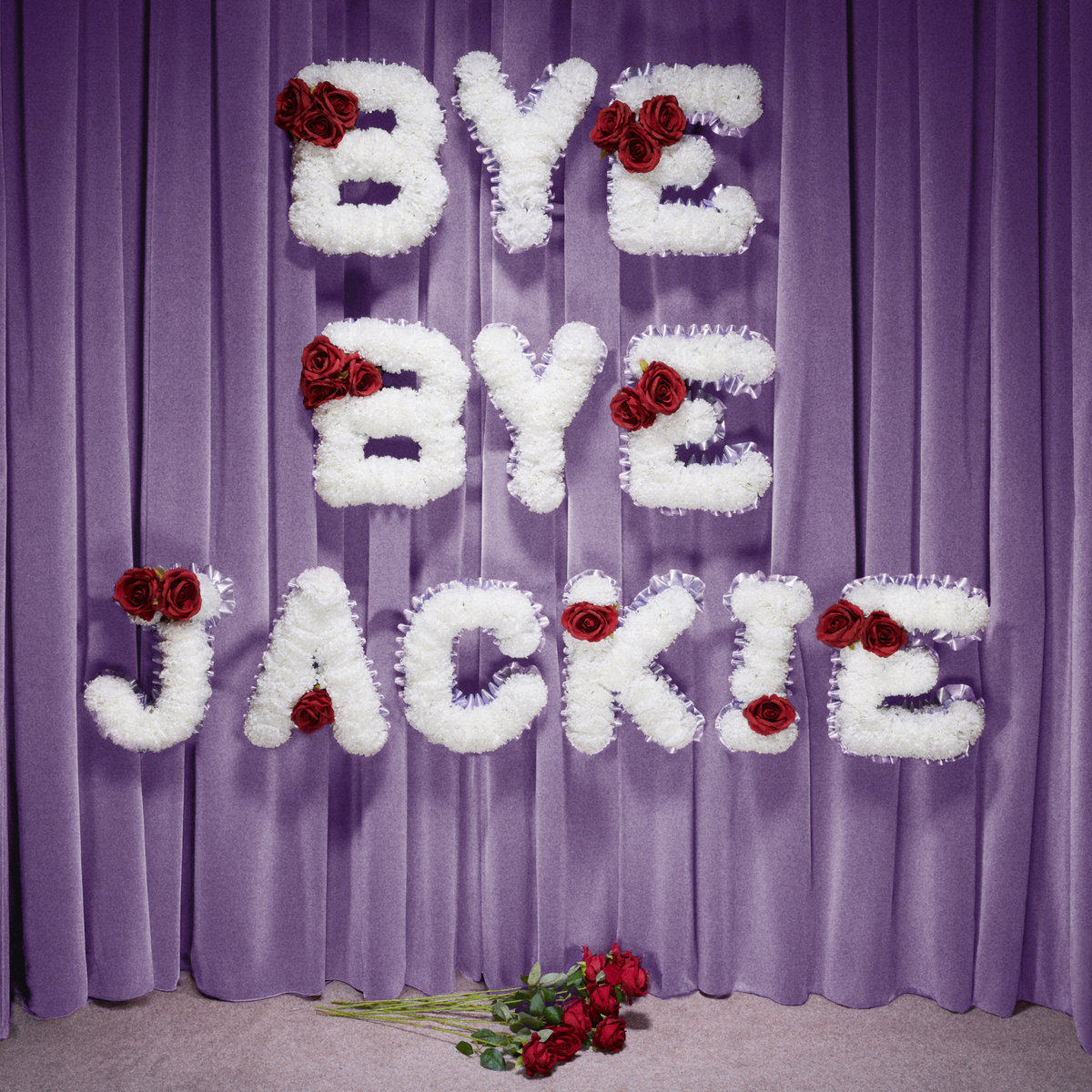 Weiße Blumenbuchstaben mit roten Rosen formen die Worte „Bye Bye Jackie“ vor einem lilafarbenen Vorhang, darunter ein Strauß roter Rosen.
