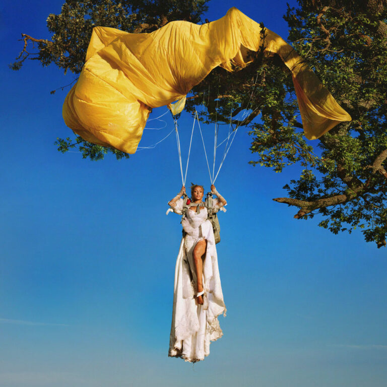 Eine Frau in weißem Kleid hängt unter einem gelben Fallschirm vor strahlend blauem Himmel, seitlich ragt ein Baum ins Bild.