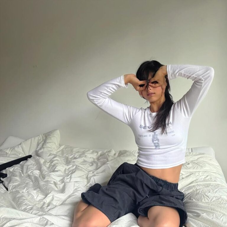 Eine junge Frau in weißem Longsleeve und dunklen Shorts sitzt auf einem ungemachten Bett, Arme über dem Kopf, Blick trotzig, schlichtes Zimmer ohne Ablenkung.