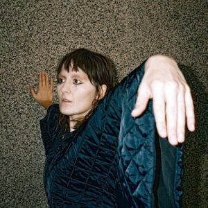 Cate Le Bon lehnt in dunklem, gemustertem Stoff an einer grauen Wand, ein Arm erhoben, der Körper in ungewöhnlicher, verdrehter Pose.