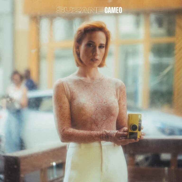 Eine Frau mit kurzem rötlichem Haar steht hinter einer Fensterscheibe, trägt ein transparentes Spitzentop und weiße Hose, hält eine gelbe Kamera in den Händen und blickt ernst nach vorn; das Bild wirkt warm, leicht unscharf und intim.