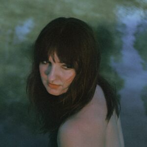 Eine junge Frau mit dunklem Haar sitzt halb im Wasser, wendet sich leicht über die Schulter und blickt in die Kamera; weiches Licht und grünlich-blauer Schimmer verleihen der Szene eine melancholische, introspektive Stimmung.