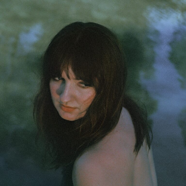 Eine junge Frau mit dunklem Haar sitzt halb im Wasser, wendet sich leicht über die Schulter und blickt in die Kamera; weiches Licht und grünlich-blauer Schimmer verleihen der Szene eine melancholische, introspektive Stimmung.
