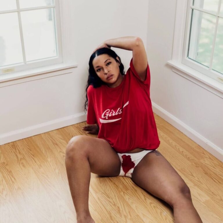 Princess Nokia sitzt in einem hellen Raum auf dem Boden, trägt ein rotes T-Shirt und weiße Unterwäsche mit sichtbarem Blutfleck – ein starkes, provokantes Motiv, das Weiblichkeit, Tabu und Selbstbestimmung in einem Bild vereint.