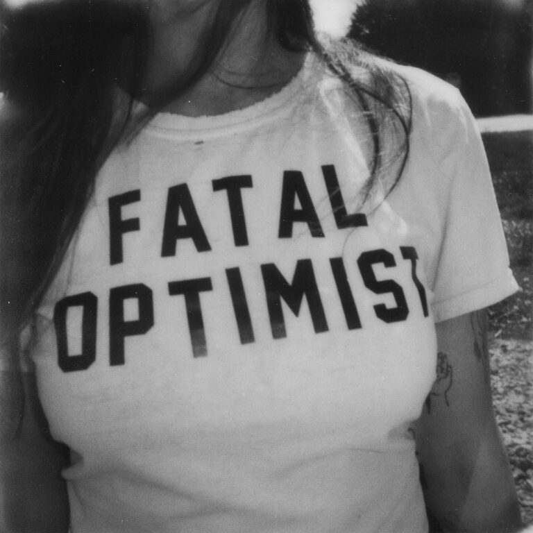 Schwarz-weißes Albumcover mit dem Schriftzug „FATAL OPTIMIST“ auf einem schlichten T-Shirt, getragen von einer Frau mit offenem Haar; Symbol für Verletzlichkeit, Klarheit und die radikale Intimität des Albums von Madi Diaz.