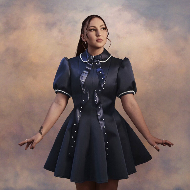 Eine Frau mit dunklem, glänzendem Kleid steht vor einem wolkigen Hintergrund, leicht seitlich gedreht, Arme ausgestreckt, der Blick ruhig und konzentriert.