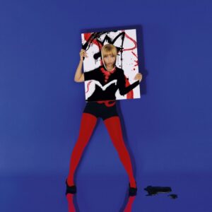 PinkPantheress steht vor tiefblauem Hintergrund, trägt schwarze Kleidung mit roten Akzenten und hält ein bemaltes Leinwandbild in den Händen. Vor ihr liegt schwarze Farbe am Boden.