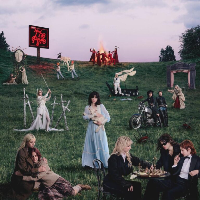 Die fünf Musikerinnen von The Last Dinner Party posieren auf einer grünen Wiese in surrealer Szenerie: Figuren mit Schwertern, Lämmern und Feuer im Hintergrund, Symbolbild für das dramatische Konzeptalbum „From The Pyre“.