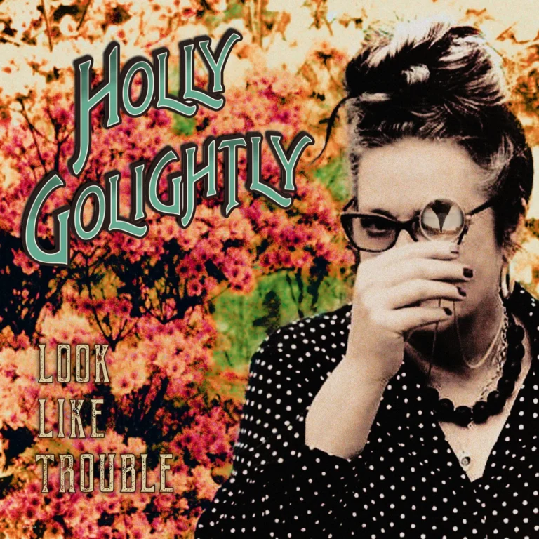 Porträt von Holly Golightly vor einem Hintergrund aus orangefarbenen Blüten, sie trägt eine gepunktete Bluse, hält ein Monokel vor das Gesicht, der Albumtitel Look Like Trouble ist in verspielter Schrift darübergelegt.
