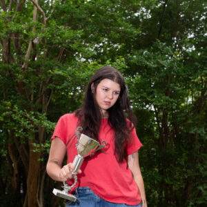 Eine junge Frau steht im Freien vor dichten grünen Bäumen, trägt ein rotes T-Shirt und Jeans, hält einen silbernen Pokal in der Hand und blickt ernst zur Seite.