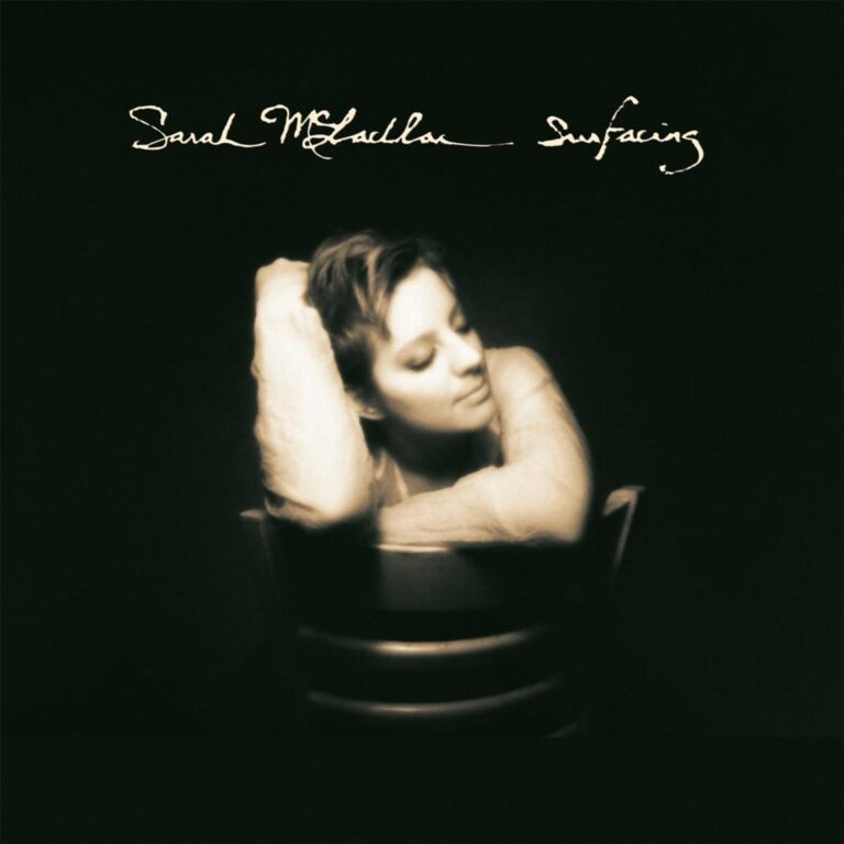 Ein monochromes, sepiafarbenes Porträt zeigt Sarah McLachlan nachdenklich auf einem Stuhl sitzend, den Kopf auf die Arme gelegt, umgeben von Dunkelheit und sanftem Lichtschein.
