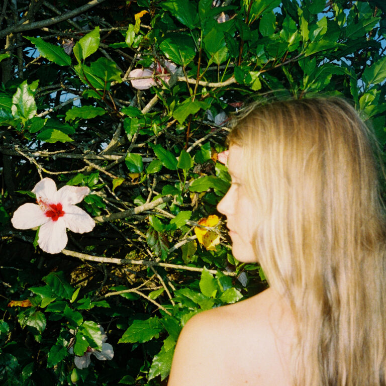 Eine blonde Frau steht seitlich vor einem grünen Strauch mit großen rosa Hibiskusblüten, ihr Blick ist ruhig und nach innen gerichtet, Licht fällt weich auf Haut und Blätter.