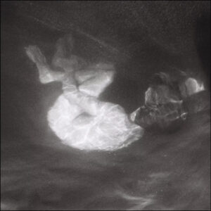 Schwarzweißfotografie zeigt eine Person unter Wasser, die in sich zusammengerollt treibt, von Lichtreflexen umhüllt, als befände sie sich zwischen Schlaf und Versinken.