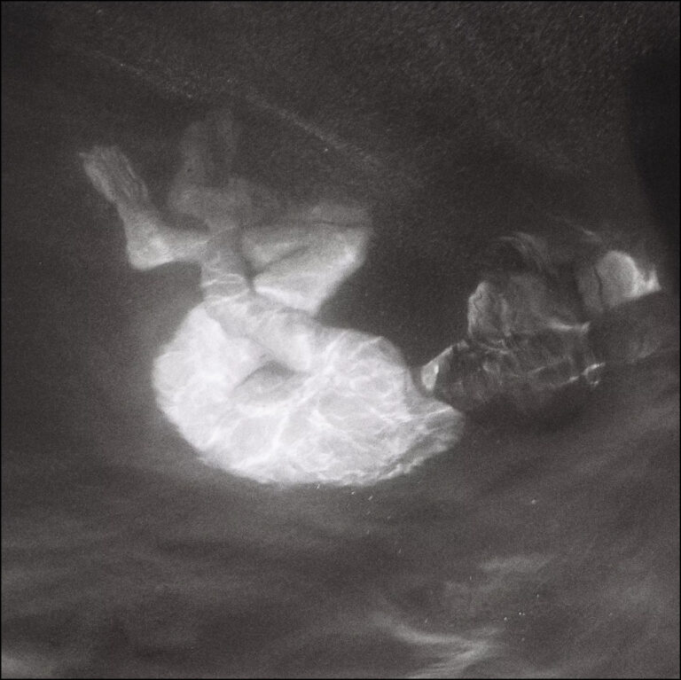 Schwarzweißfotografie zeigt eine Person unter Wasser, die in sich zusammengerollt treibt, von Lichtreflexen umhüllt, als befände sie sich zwischen Schlaf und Versinken.