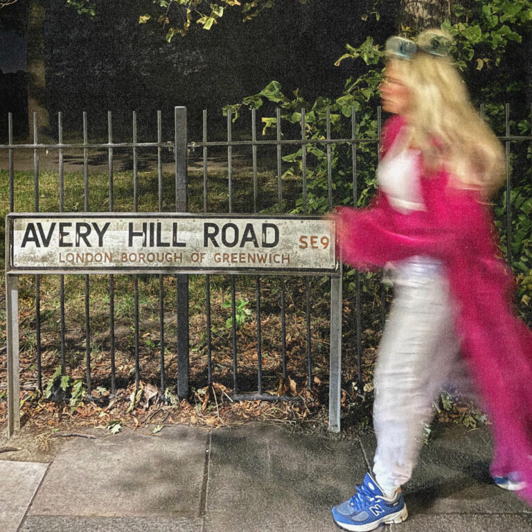 Eine blonde Frau in Bewegung vor einem Straßenschild mit der Aufschrift „Avery Hill Road – London Borough of Greenwich“. Nachtaufnahme mit urbaner, atmosphärischer Beleuchtung.