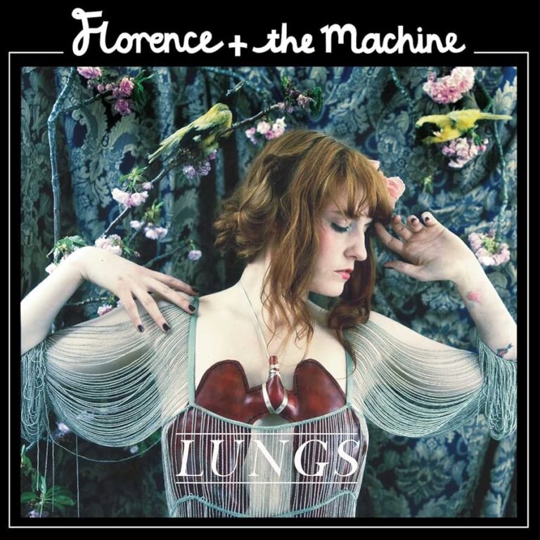 Florence Welch in transparentem Kleid mit künstlichen Lungen auf der Brust, umgeben von dunklem Blumenschmuck und schimmernden Stoffen – zwischen Anmut und Bedrängnis.