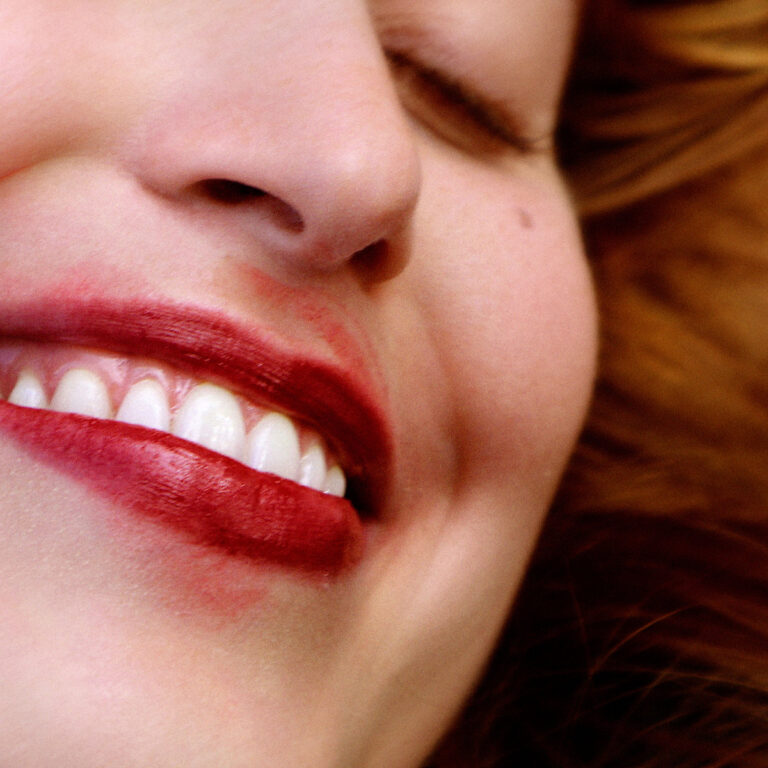Nahaufnahme eines lachenden Gesichts mit verwischtem rotem Lippenstift, halb geschlossene Augen, warmer Hautton, rotes Haar im Hintergrund, spontane Momentaufnahme mit sinnlicher Unschärfe.