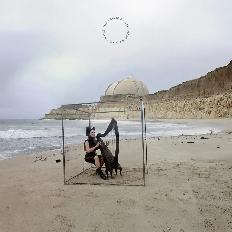 Allie X sitzt auf einem leeren Strand in einem transparenten Würfel und spielt eine schwarze Harfe, während hinter ihr eine futuristische Kuppel aus dem Sand ragt und der Himmel milchig grau über dem Meer hängt.
