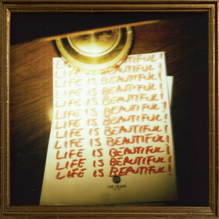Ein vergilbter Zettel liegt auf einem Holztisch unter einer goldenen Lampe, darauf in roter Schrift mehrfach der Satz „LIFE IS BEAUTIFUL!“. Das Bild wirkt wie ein manisches Bekenntnis zwischen Verzweiflung und Hoffnung.