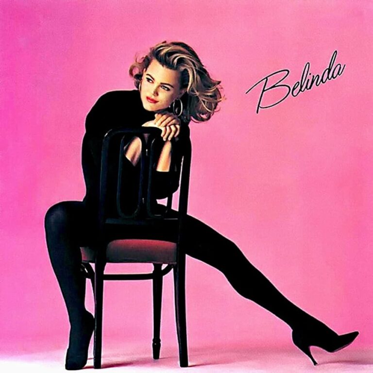 Belinda Carlisle sitzt in schwarzem Outfit auf einem Stuhl vor rosafarbenem Hintergrund, der Körper angespannt, der Blick konzentriert in die Ferne gerichtet.