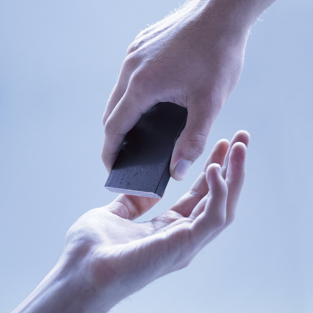 Zwei Hände in kaltem Licht. Eine Hand reicht ein dunkles rechteckiges Objekt an eine andere weiter. Die Szene wirkt distanziert und minimalistisch.