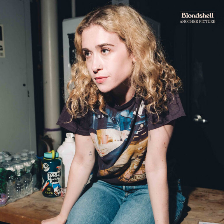 Eine junge Frau mit blonden Locken sitzt in einem schlichten Raum auf einer Holzfläche, stützt ihre Arme auf und blickt ernst zur Seite. Helles Licht fällt auf ihr Gesicht, im Hintergrund stehen Wasserflaschen, ein Getränkebehälter und ein dunkler Türrahmen. Die Atmosphäre wirkt ruhig und nachdenklich.