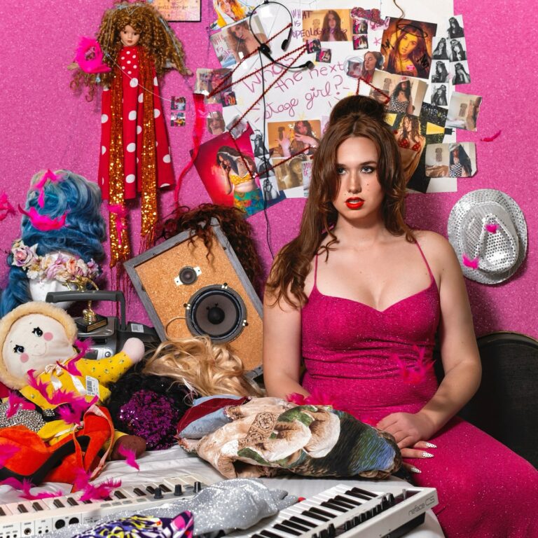 Eine junge Frau sitzt in einem überfüllten pinken Zimmer voller Perücken, Puppen, Fotos und Requisiten. Sie trägt ein glitzerndes pinkes Kleid und blickt ernst in die Kamera. Um sie herum liegen ein Keyboard, Federn, Stoffpuppen und Collagen aus Popbildern.