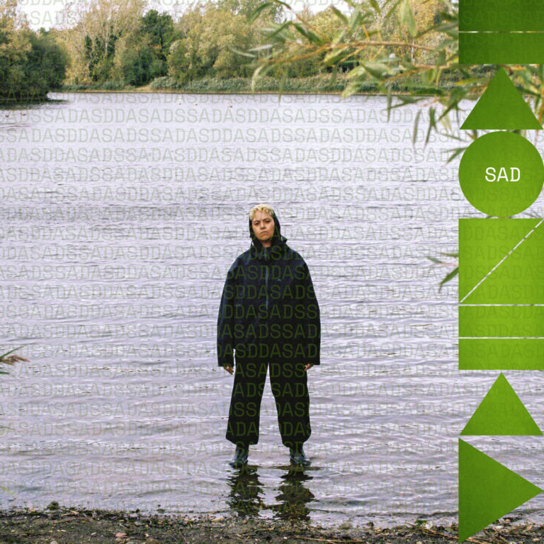 Eine Person in dunkler Kleidung steht im flachen Wasser eines Sees, umgeben von herbstlichen Bäumen. Rechts im Bild befinden sich grüne grafische Formen und der Schriftzug SAD.