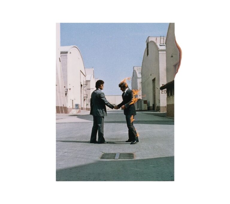 Zwei Männer in grauen Anzügen reichen sich auf einem Filmstudiogelände die Hand, während einer der beiden in Flammen steht. Die Gebäude werfen klare Schatten und der helle Himmel bildet einen ruhigen Hintergrund.