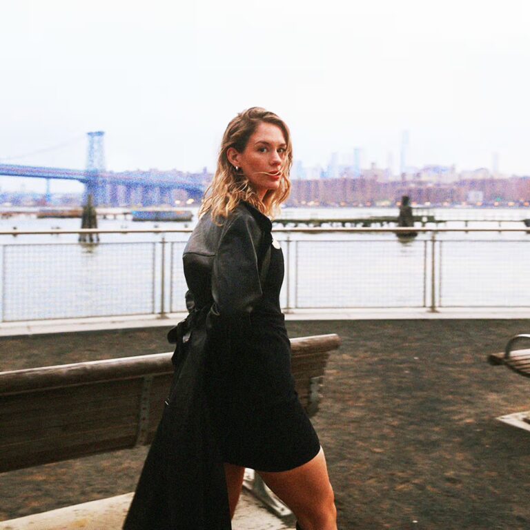 Eine Frau steht an einer Uferpromenade, trägt ein dunkles Kleid, dreht sich halb zur Kamera, im Hintergrund Wasser, Brücke, verwaschene Stadtsilhouette im blassen Licht.