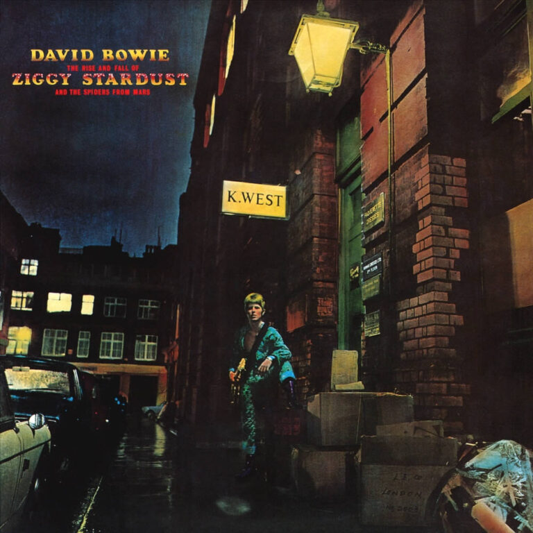 David Bowie steht nachts auf einer Londoner Straße unter einem leuchtenden Schild „K. West“, nasses Pflaster und dunkle Fassaden im Laternenlicht.