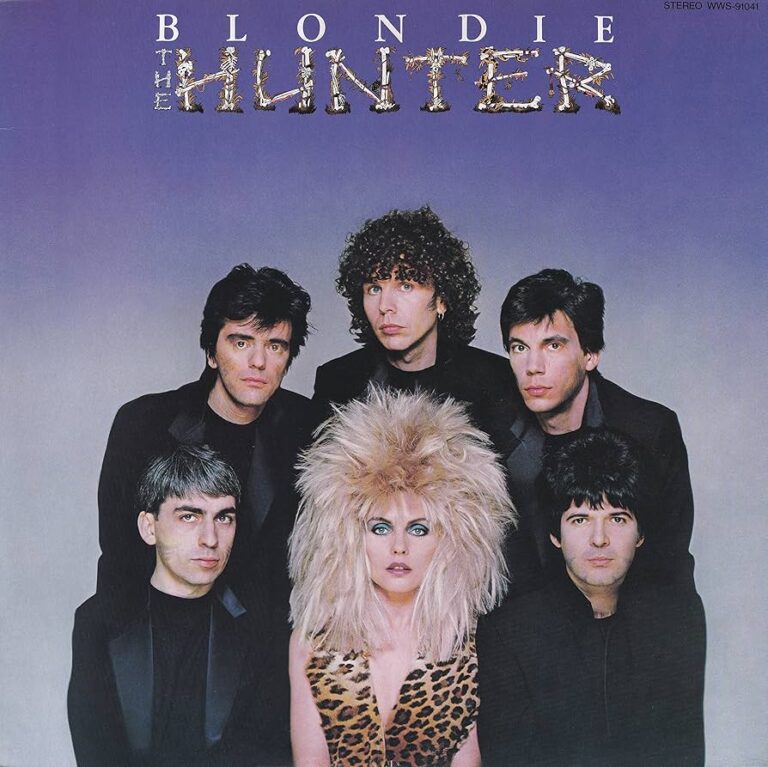 Blondie auf dem Albumcover von „The Hunter“: Debbie Harry frontal inszeniert mit voluminöser Frisur, die Band im Hintergrund, dunkler Studiokontext, kontrollierte Pose und direkter Blick.