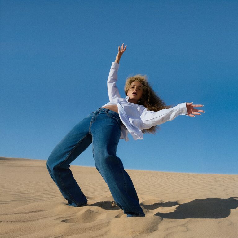 Eine Frau steht in einer Dünenlandschaft, lehnt sich weit nach hinten, trägt Jeans und ein weißes Hemd, der Himmel leuchtet tiefblau.