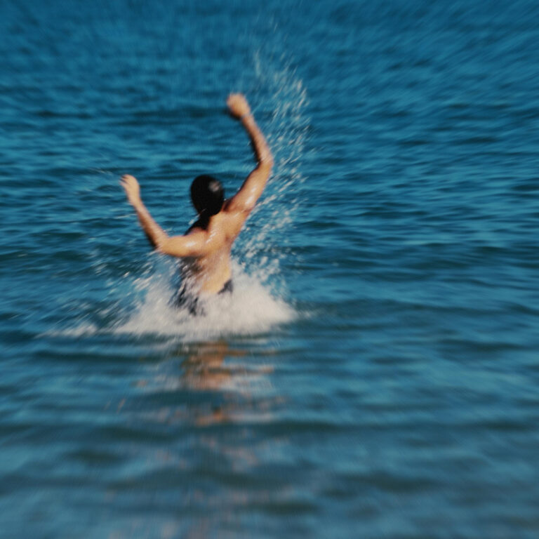 Eine unscharfe Aufnahme zeigt eine Person von hinten, die mit erhobenen Armen ins offene Wasser springt. Die Bewegung wirkt impulsiv, das Wasser spritzt auf, der Horizont bleibt diffus und weit.
