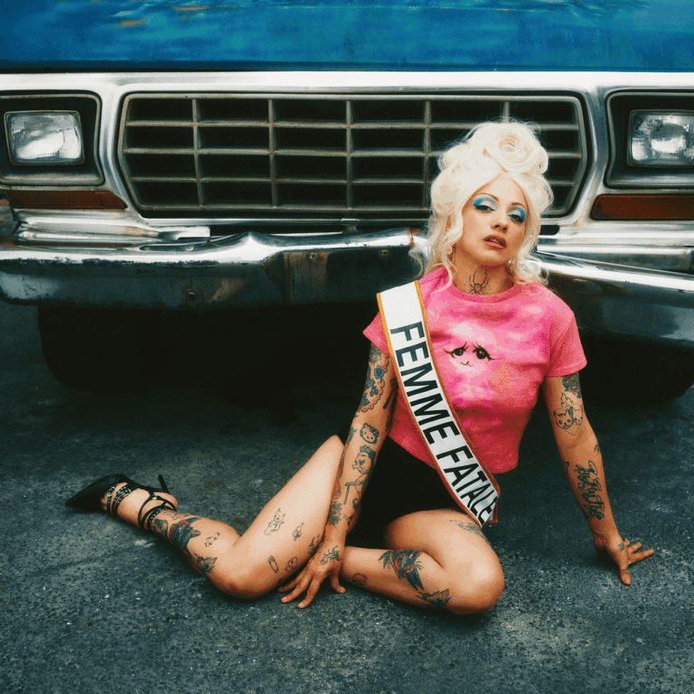 Mon Laferte sitzt im pinken Kleid auf dem Boden vor einem Auto, trägt eine Schärpe mit der Aufschrift Femme Fatale, das Bild wirkt inszeniert und verletzlich zugleich.