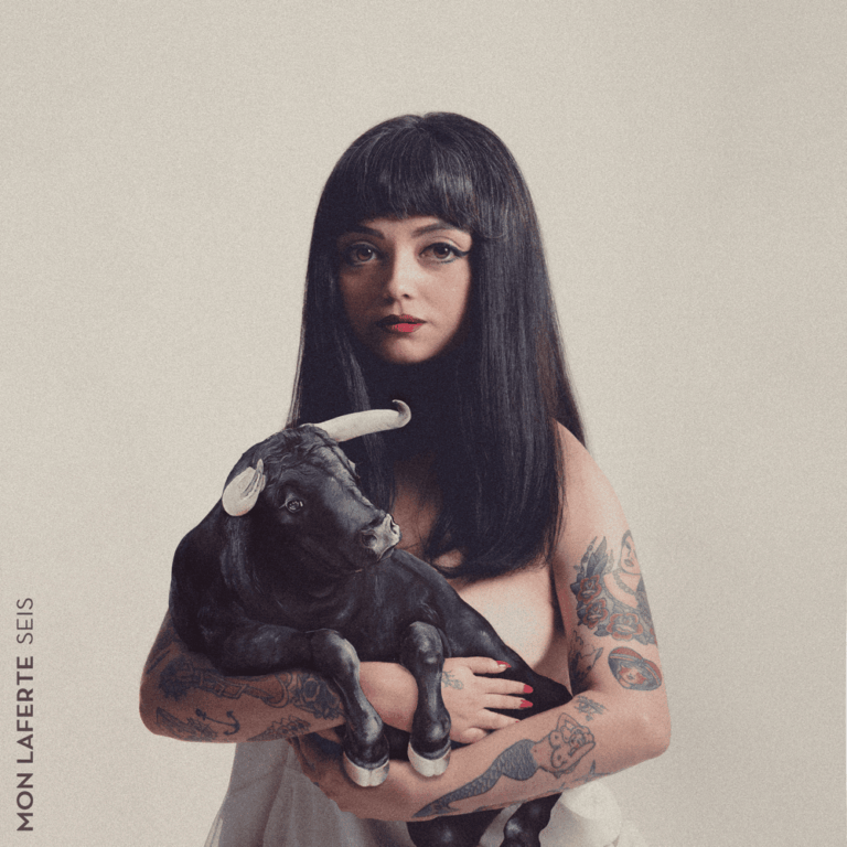 Porträt von Mon Laferte vor hellem Hintergrund, sie hält einen schwarzen Hund im Arm, direkter Blick, reduzierte Farbgebung, ruhige Bildkomposition.
