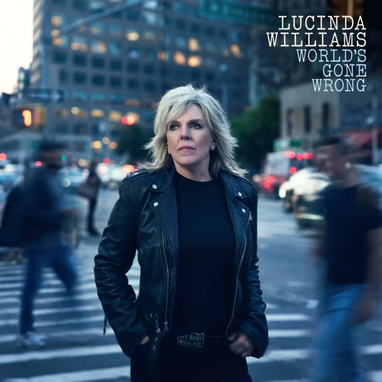 Porträt von Lucinda Williams in dunkler Kleidung auf einer belebten Stadtstraße, umgeben von unscharfen Passanten, ernst und fokussiert blickend.