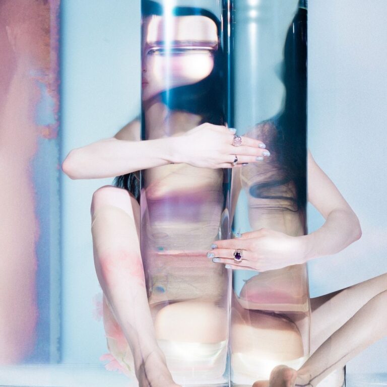 Abstrakt inszeniertes Porträt einer Frau hinter transparenten Glasflächen, fragmentiert und spiegelnd, mit kühler Farbpalette und kontrollierter Körperhaltung.