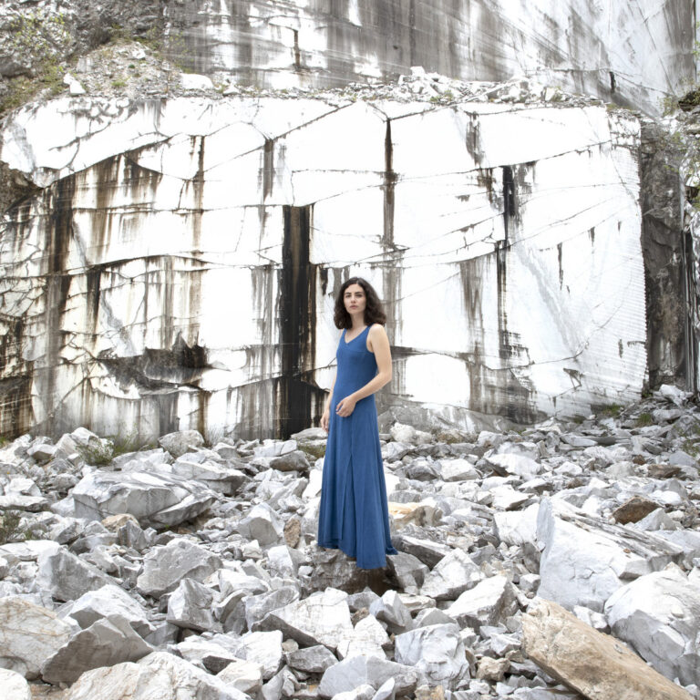 Marta Del Grandi steht in einem blauen Kleid in einer felsigen, hellen Steinlandschaft vor einer steilen Felswand, ruhig und nachdenklich inszeniert.