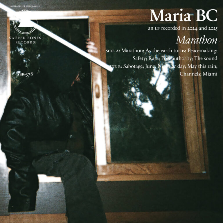 Maria BC sitzt teilweise verdeckt vor einem Fenster, Reflexionen überlagern das Gesicht, dunkle Töne dominieren das Bild.