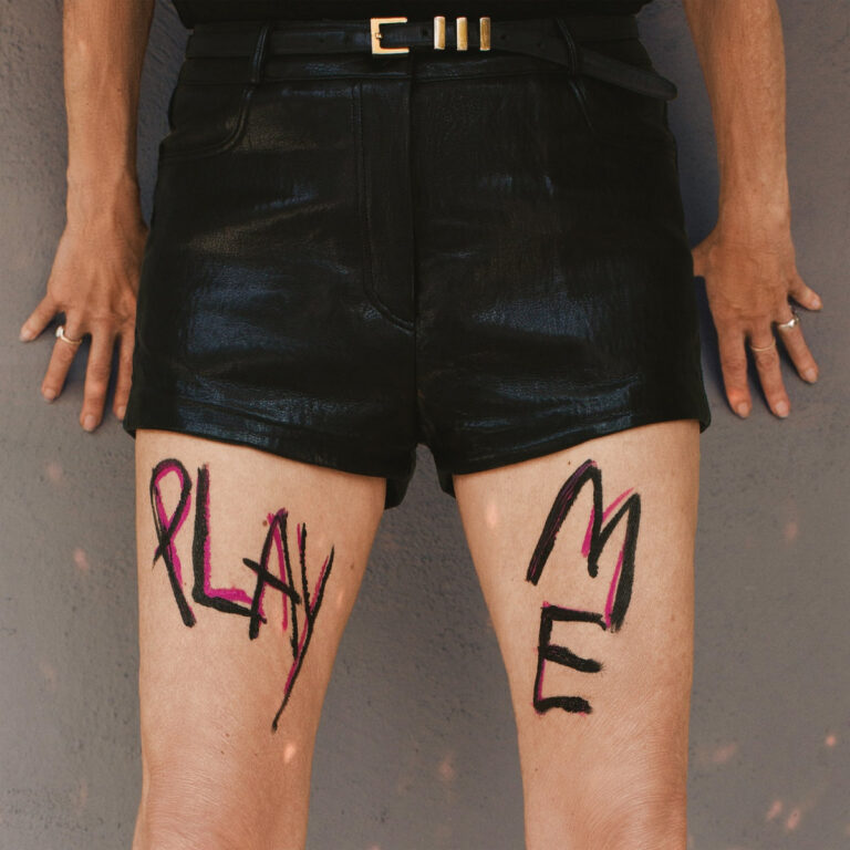 Nahaufnahme der Beine und des Unterkörpers einer Person in schwarzen Ledershorts vor einer grauen Wand, wobei die Worte „PLAY“ und „ME“ in schwarzer und pinker Farbe großflächig auf die Oberschenkel gemalt sind.