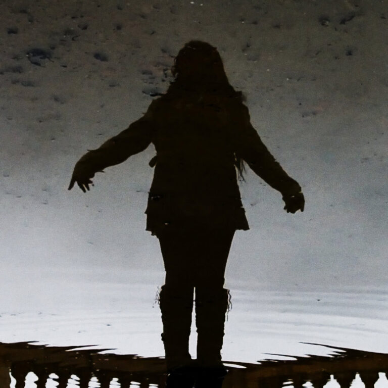 Eine dunkle Silhouette einer Person mit ausgebreiteten Armen spiegelt sich in einer leicht bewegten, dunklen Wasserfläche. Der Hintergrund ist körnig und grau-braun meliert, was eine düstere, melancholische Atmosphäre erzeugt.