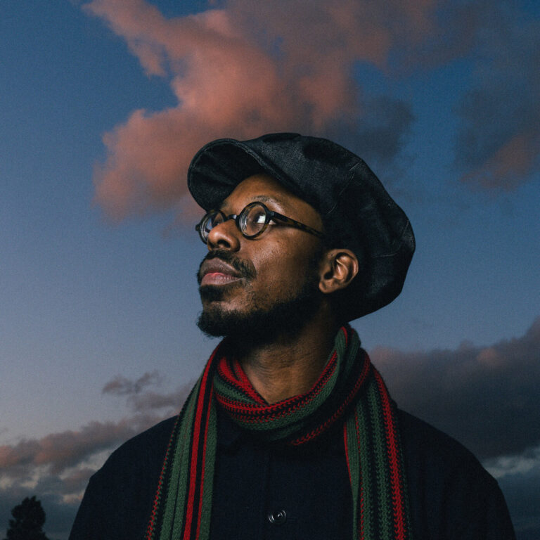 Porträt von Shabaka vor dämmrigem Wolkenhimmel, nach oben blickend, mit Schiebermütze, Brille sowie einem Schal in panafrikanischen Farben.