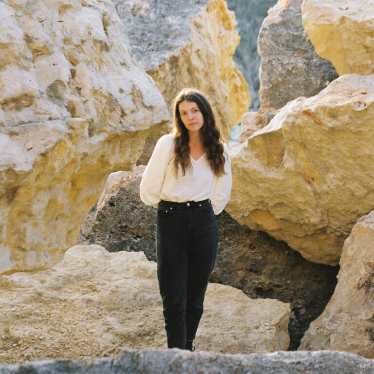 Singer-Songwriterin Natalie Jane Hill steht in weißer Bluse und dunkler Hose zentral zwischen hellen, zerklüfteten Felsformationen und blickt direkt in die Kamera.