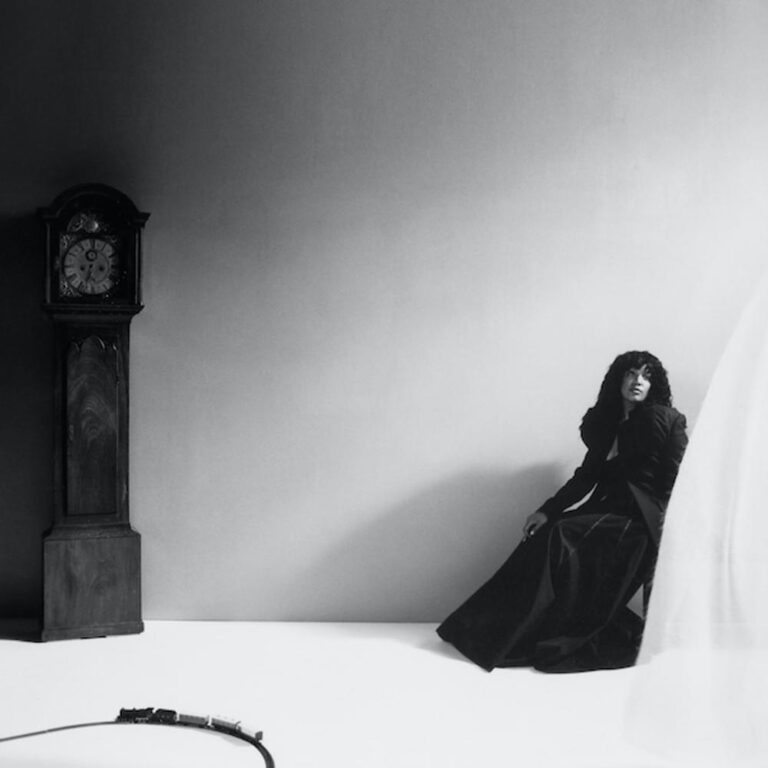 Schwarz-Weiß-Fotografie von Arima Ederra, die in einem langen dunklen Kleid in einer Raumecke neben einer antiken Standuhr sitzt, umgeben von tiefen Schatten sowie weichem, diffusem Licht.