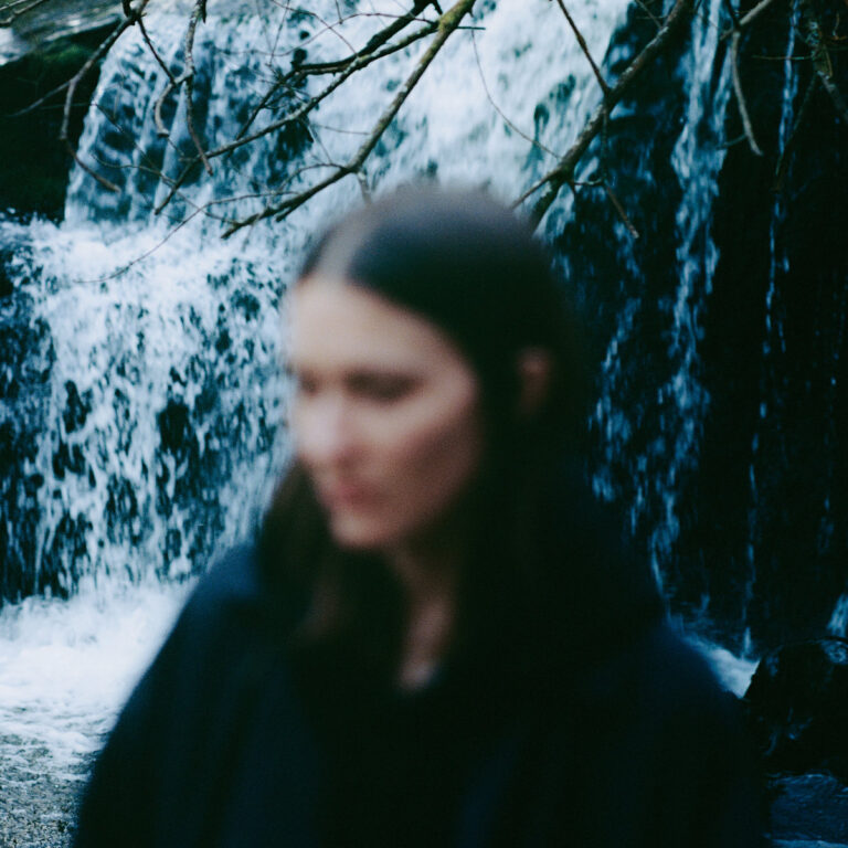 Verschwommenes Porträt von Polly Mackey vor einem Wasserfall im Wald, kühle Blautöne dominieren die Szenerie.