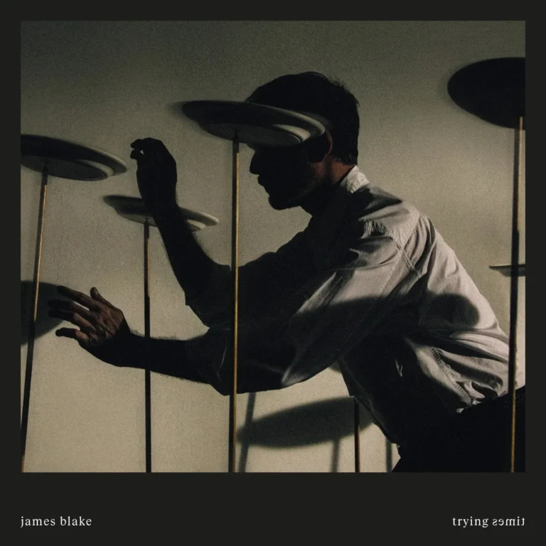 James Blake als dunkle Silhouette vor einer hellen Kulisse, während er mehrere auf Stäben balancierte Teller mit erhobenen Händen in Bewegung hält.