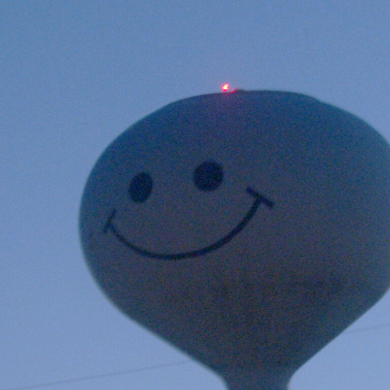 Ein großer, hellgrauer Wasserturm in Form eines runden Kopfes mit einem aufgemalten, simplen schwarzen Smiley-Gesicht steht vor einem dämmrig blauen Abendhimmel. Auf der Spitze des Kopfes leuchtet ein kleiner, roter Punkt.