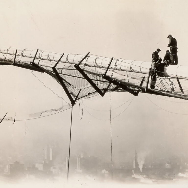 Eine historische Schwarz-Weiß-Fotografie zeigt vier Bauarbeiter in schwindelerregender Höhe auf einem schmalen Metallgerüst einer Brückenkonstruktion über San Francisco. Unter ihnen erstreckt sich eine im Nebel versinkende Stadtlandschaft, während sie an einem massiven, mit Seilen umwickelten Kabel arbeiten.